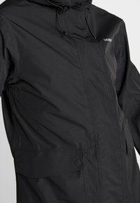 Veste imperméable noire avec fermeture éclair avant, capuche et poches à boutons-pression. Comprend des poignets élastiques et des cordons de serrage ajustables.