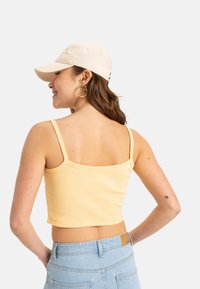 Gul ribbet crop top med tynde stropper, parret med lyseblå højtaljede jeans. Modellen bærer en beige baseballcap og hoops øreringe.