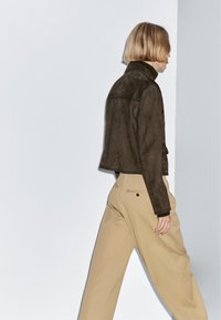 Hnědá bledohnědá krátká semišová bunda s vysokým límcem, doplněná světle béžovými širokými kalhotami. Jednoduchý, minimalistický design, neutrální barevná paleta.