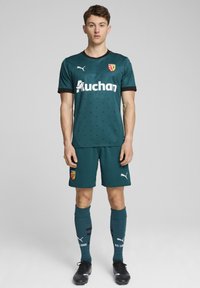 Grön fotbollströja i andningsbart material med korta ärmar, svart trim, logotyp och mönstrade detaljer. Kompletteras med matchande shorts och strumpor.