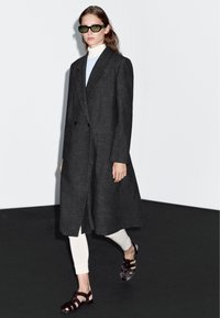 Dlouhý tmavě šedý kabát s klopovým límcem, dvouřadým zapínáním, nošený přes světle modrý rolák, doplněný bílými legínami a hnědými sandály.