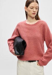 Roza pletena sv sweater z okroglim izrezom, rebrastimi manšetami in subtilnimi diagonalnimi vzorci. Kombinirana z črno teksturirano torbico in smetanastimi hlačami.