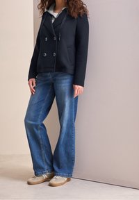 Femme portant une veste noire croisée sur un sweat à capuche gris, un jean bleu à jambes larges et des baskets beige, debout contre un mur pâle.