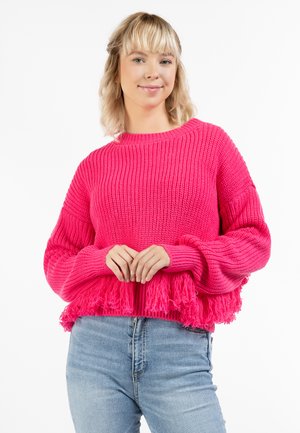 Felroze gebreide trui met een textuur ribbelpatroon, wijde mouwen en een franjenzoom. Gecombineerd met lichtblauwe high-waisted jeans.