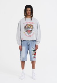 Felpa grigia con grafica di tigre e testo "Ed Hardy"; pantaloni corti di jeans con disegni colorati in stile tatuaggio. Sneakers bianche e calze corte completano il look.
