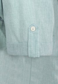 Manga de camisa de tela azul claro que cuenta con un cierre de botón único, puño puntiagudo y un acabado suave y texturizado. Costuras mínimas visibles.