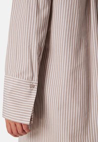 Camicia beige a maniche lunghe con righe bianche verticali, dotata di un bottone decorativo sul polsino e realizzata in morbido cotone.