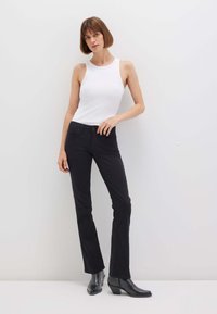 Donna con capelli castani corti che indossa una canotta bianca senza maniche, jeans neri aderenti e stivali neri alla caviglia, in piedi contro un muro bianco semplice.