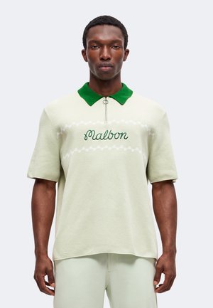 Mann trägt hellgrünes kurzärmeliges Poloshirt mit grünem Kragen und "Malbon" auf der Brust gestickt, steht vor einem einfarbigen Hintergrund.
