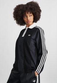 Pulover sport negru cu guler alb, textură cu model dantelat pe față și trei dungi albe pe fiecare mânecă.