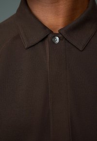 Braunes, strukturiertes Poloshirt mit einem gewebten Muster, das einen spitzen Kragen und einen einzelnen Knopfverschluss am Hals hat.