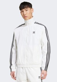 Vit zip-jacka med hög krage, svarta ränder längs ärmarna, elastiska cuffs och midja. Har Adidas-logotyp på bröstet.