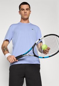 Ανοιχτό μπλε αθλητικό μπλουζάκι με κοντά μανίκια και πράσινο λογότυπο, συνδυασμένο με μαύρα αθλητικά παντελόνια. Κρατώντας μια ρακέτα τένις και μια μπάλα στα χέρια.