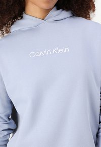 Svetlo modra kapuca z mehkimi vlakni, z belim logotipom "Calvin Klein" spredaj in klasičnim dizajnom s kolebnico za nastavitev kapuce.