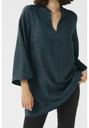 Kvinde iført mørk teal textured bluse med lange ærmer og flæset V-hals, kombineret med sorte bukser, holder blusens side.