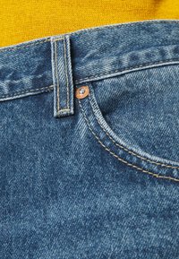 Vaqueros de mezclilla en azul medio con costuras visibles; presentan un remache de cobre y una abertura de bolsillo frontal, tejido texturizado y un pasador para cinturón.