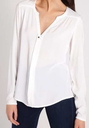 Blouse blanche à manches longues avec un col en V, patte de boutonnage sur le devant, tissu doux et ourlet droit. Associée à un denim foncé.