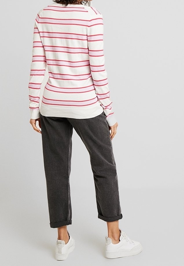 Femme portant un pull blanc à rayures horizontales rouges, un jean foncé retroussé et des baskets blanches, debout de dos sur un fond uni.