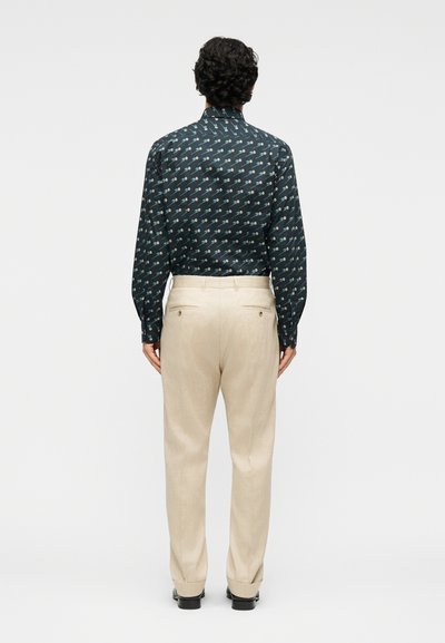 Άνδρας που στέκεται με την πλάτη προς τα εμπρός, φοράει σκούρο πουκάμισο με μοτίβο και μακριά μανίκια, το οποίο είναι βιδωμένο μέσα σε μπεζ παντελόνι, και μαύρα παπούτσια, σε απλό φόντο.