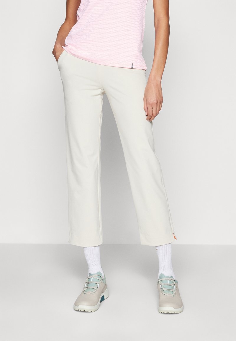 Personne portant un pantalon court écru, des chaussettes blanches, des baskets beiges et un haut sans manches rose clair, debout avec une main dans la poche.