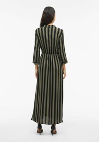 Dlhé priliehavé šaty v olivovo zelenej a čiernych vertikálnych prúžkoch, s trojštvrťovými rukávmi a elastickým pásom, doplnené čiernymi lodičkami.