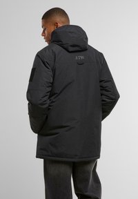 Černá zateplená bunda s kapucí, která má texturovaný povrch, nastavitelné šňůrky a logo na zadní straně. Vyztužené záplaty na loktech.