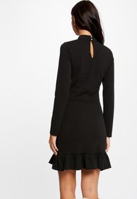 Vestido negro de manga larga con dobladillo con volantes, detalle de abertura en la parte posterior y tela texturizada, presentando un cuerpo ajustado y una falda acampanada.