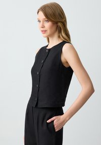 Top corto senza maniche di colore nero con scollatura rotonda, caratterizzato da sei bottoni frontali e una tasca sul lato sinistro, realizzato in tessuto testurizzato.
