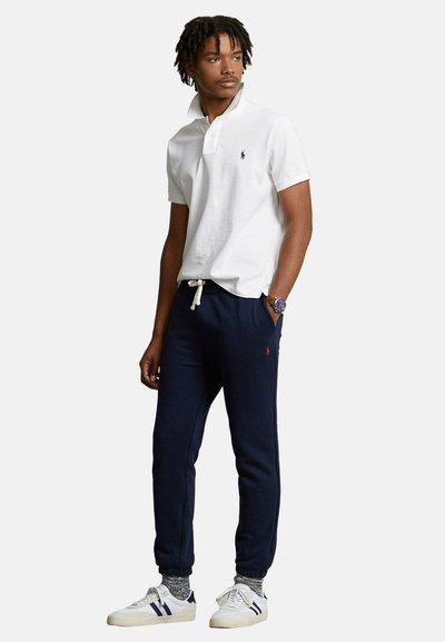 Uomo con dreadlocks che indossa una polo bianca, pantaloni della tuta blu navy, calzini grigi e scarpe da ginnastica bianche, in piedi con una mano in tasca.