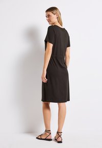 Musta istuva mekko lyhyillä hihoilla, vyötäröltä kiristetty ja sileää kangasta. Yhdistetty mustiin hihnakenkiin. Takaview on esillä.