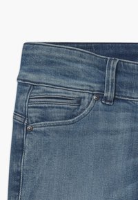 Modré džíny z denimu mají klasický pětikapsový design, s barevným šitím a mosaznými akcenty na pase a kapsách.