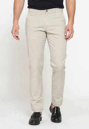 Uomo che indossa pantaloni beige chiaro slim fit e scarpe slip-on in pelle nera in piedi davanti a uno sfondo semplice.