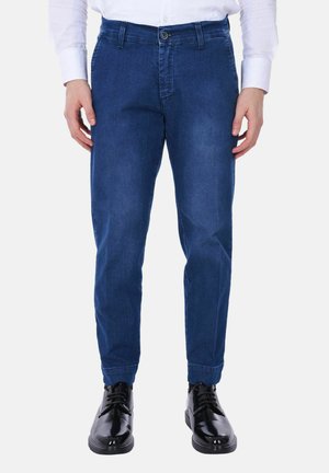Uomo che indossa jeans slim fit blu scuro, camicia bianca con bottoni e scarpe eleganti nere lucide, in piedi su sfondo uniforme.