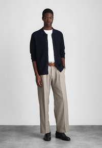 Cardigan a maglia blu navy sopra una t-shirt bianca, abbinato a pantaloni beige chiaro. Cintura in pelle marrone e scarpe nere. Design minimalista.