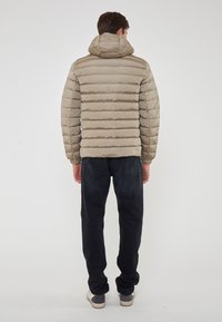 Giacca beige imbottita con cuciture orizzontali, cappuccio e polsini elastici, indossata con jeans scuri e sneaker. Sfondo bianco semplice.