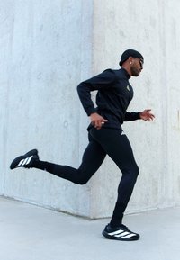 Uomo che indossa abbigliamento sportivo nero e occhiali da sole corre accanto a un muro di cemento su una superficie lastricata.