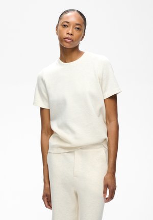 Femme aux cheveux attachés portant un t-shirt blanc uni à manches courtes et un pantalon blanc assorti sur fond blanc.