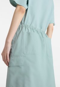 Light blue, elastic-waisted dress with short sleeves, featuring two front patch pockets and a smooth, textured fabric finish.