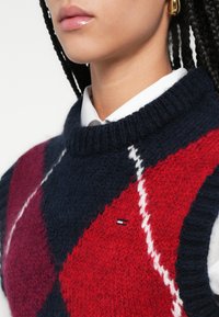 Pletený sveter so vzorom argyle v námorníckej modrej, červenej a bordovej farbe. Rebrovaný okrúhly výstrih a logo na hrudi. Mäkká textúra.