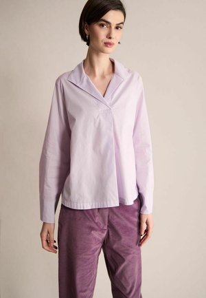 Femme aux cheveux courts et foncés portant un chemisier à manches longues couleur lavande claire et un pantalon en velours côtelé violet, debout devant un fond neutre.