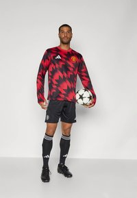 Maglia da portiere rossa e nera con motivi geometrici, con lo stemma del Manchester United, abbinata a pantaloncini e calzettoni neri. Tiene in mano un pallone da calcio.