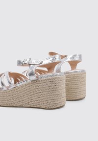 Sandalias metálicas plateadas con tiras y tacón de cuña de esparto, que presentan una superficie texturizada y una correa ajustable en el tobillo.