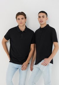 Due persone indossano polo nere a maniche corte in cotone, abbinate a jeans azzurri chiari. Il design presenta un colletto classico con bottoni.