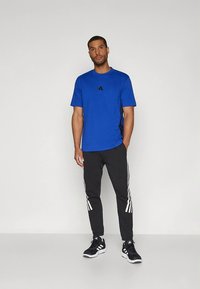 Μπλε αθλητική κοντομάνικη μπλούζα με μαύρο λογότυπο, συνδυασμένη με μαύρο παντελόνι φόρμας με λευκές ρίγες στα πλάγια και μαύρα αθλητικά παπούτσια.