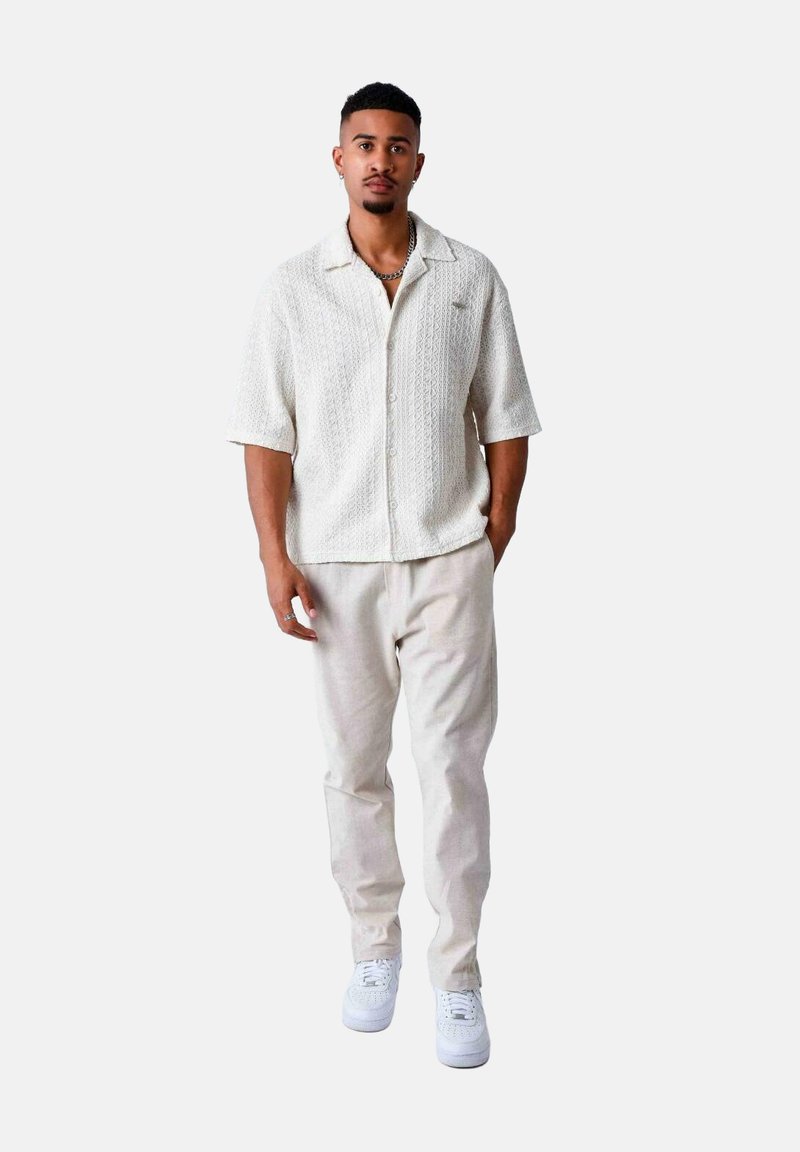 Chemise blanche en tricot à manches courtes avec un motif texturé, associée à un pantalon beige clair et des baskets blanches, se tenant devant un fond blanc.