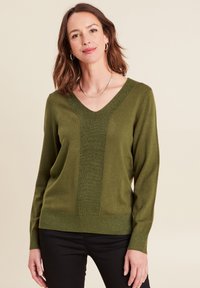 Groene gebreide trui met een V-hals, textuur in het midden, lange mouwen en een aansluitende onderkant. Gecombineerd met zwarte broeken.
