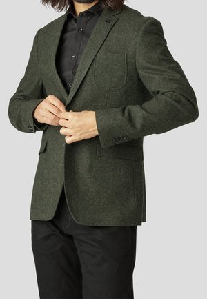 Homme boutonnant un blazer en laine vert foncé sur une chemise noire et un pantalon noir sur un fond gris clair uni.