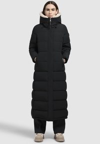 Vrouw in een lange zwarte gewatteerde jas met een hoge kraag, gevoerd met beige fleece, staand met handen in de zakken, gekleed in zwarte broek en beige schoenen.