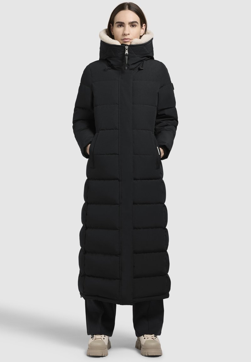 Vrouw in een lange zwarte gewatteerde jas met een hoge kraag, gevoerd met beige fleece, staand met handen in de zakken, gekleed in zwarte broek en beige schoenen.