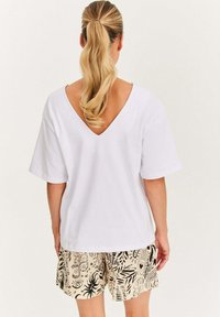 Kobieta z blond włosami związanymi w kucyk, ubrana w białą koszulkę z dekoltem w kształcie litery V i jasnobeżowe szorty z czarnymi abstrakcyjnymi wzorami, widziana z tyłu.